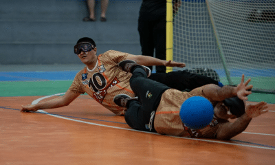 Dois jogadores de goalball em quadra indoor laranja durante uma defesa. Ambos usam vendas pretas nos olhos, camisas amarelo‑douradas com detalhes em laranja e números grandes na frente, calças escuras e tênis esportivos. O jogador mais ao fundo está deitado de lado, quase na horizontal em relação ao chão, com o braço esquerdo estendido para frente e expressão de esforço; o número 10 aparece com destaque na camisa dele. À frente, outro jogador está totalmente estendido no chão, com o corpo deitado de lado e braços abertos; uma bola azul de goalball está encostada na parte lateral do corpo ou do rosto dele, na altura da cabeça. As pernas dos dois atletas se cruzam, transmitindo a ideia de um movimento rápido e conjunto para bloquear a bola. Ao fundo, aparece a trave com rede clara e parte do piso da quadra em verde e azul, além de pernas de outras pessoas em pé próximas à área do gol.