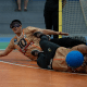 Dois jogadores de goalball em quadra indoor laranja durante uma defesa. Ambos usam vendas pretas nos olhos, camisas amarelo‑douradas com detalhes em laranja e números grandes na frente, calças escuras e tênis esportivos. O jogador mais ao fundo está deitado de lado, quase na horizontal em relação ao chão, com o braço esquerdo estendido para frente e expressão de esforço; o número 10 aparece com destaque na camisa dele. À frente, outro jogador está totalmente estendido no chão, com o corpo deitado de lado e braços abertos; uma bola azul de goalball está encostada na parte lateral do corpo ou do rosto dele, na altura da cabeça. As pernas dos dois atletas se cruzam, transmitindo a ideia de um movimento rápido e conjunto para bloquear a bola. Ao fundo, aparece a trave com rede clara e parte do piso da quadra em verde e azul, além de pernas de outras pessoas em pé próximas à área do gol.