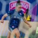 Na imagem, através das redes do gol adversário, se vê Dudinha conduzindo a bola no ataque.