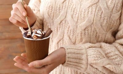 Pessoa segurando uma bebida quente ao lado de uma pequena porção de chocolate ou bolo caseiro, em ambiente aconchegante de inverno.